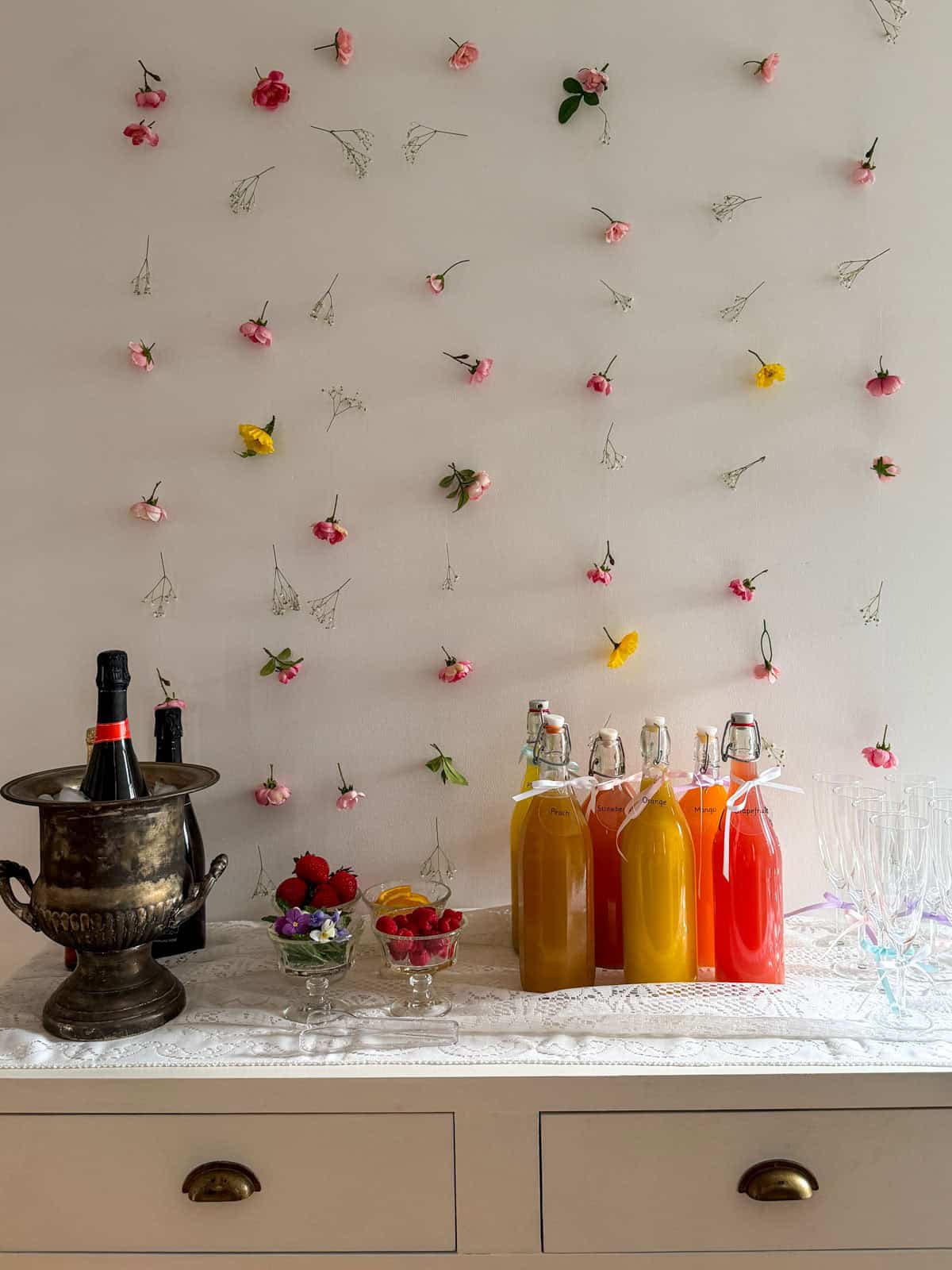 Full mimosa bar setup on white console table with champagne bucket, fruit juice bottles, garnish bowls and ribbon-tied flutes against white wall with scattered pink and yellow flowers.