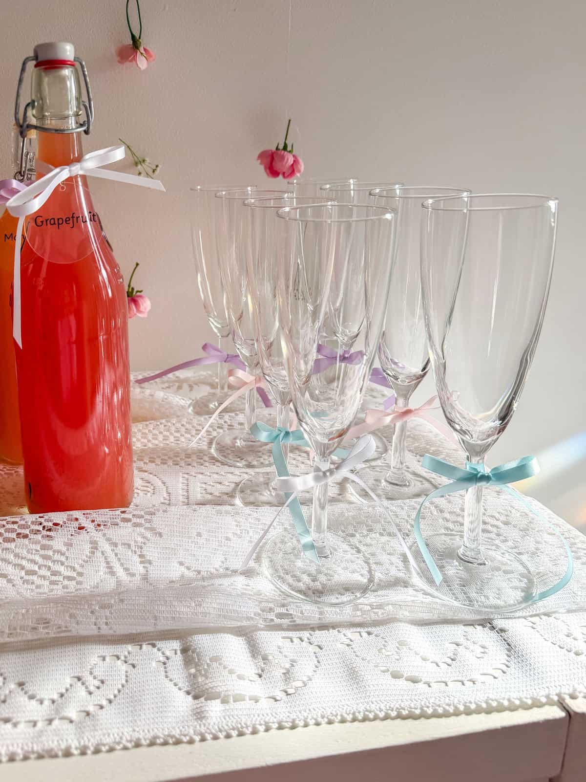 Row of crystal champagne flutes with pastel satin ribbon bows beside a grapefruit juice bottle with pink flowers on white wall behind.