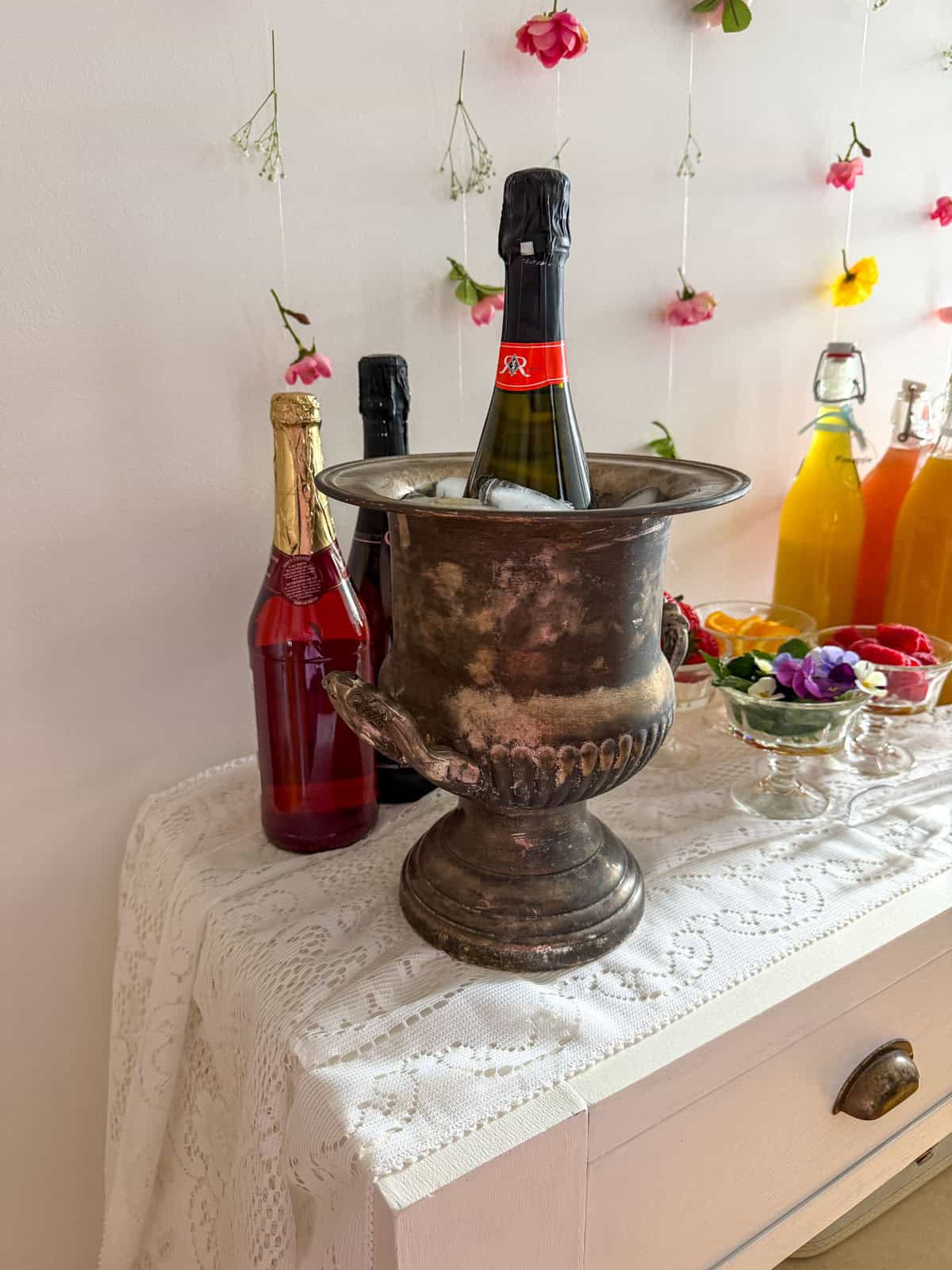 Vintage silver champagne bucket with sparkling wine bottle on white lace tablecloth with colorful juice bottles and hanging flowers in background.