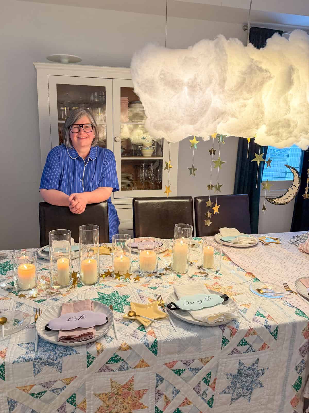 Hostess in blue striped pajamas standing at a breakfast for dinner party table with cloud and star installation overhead.