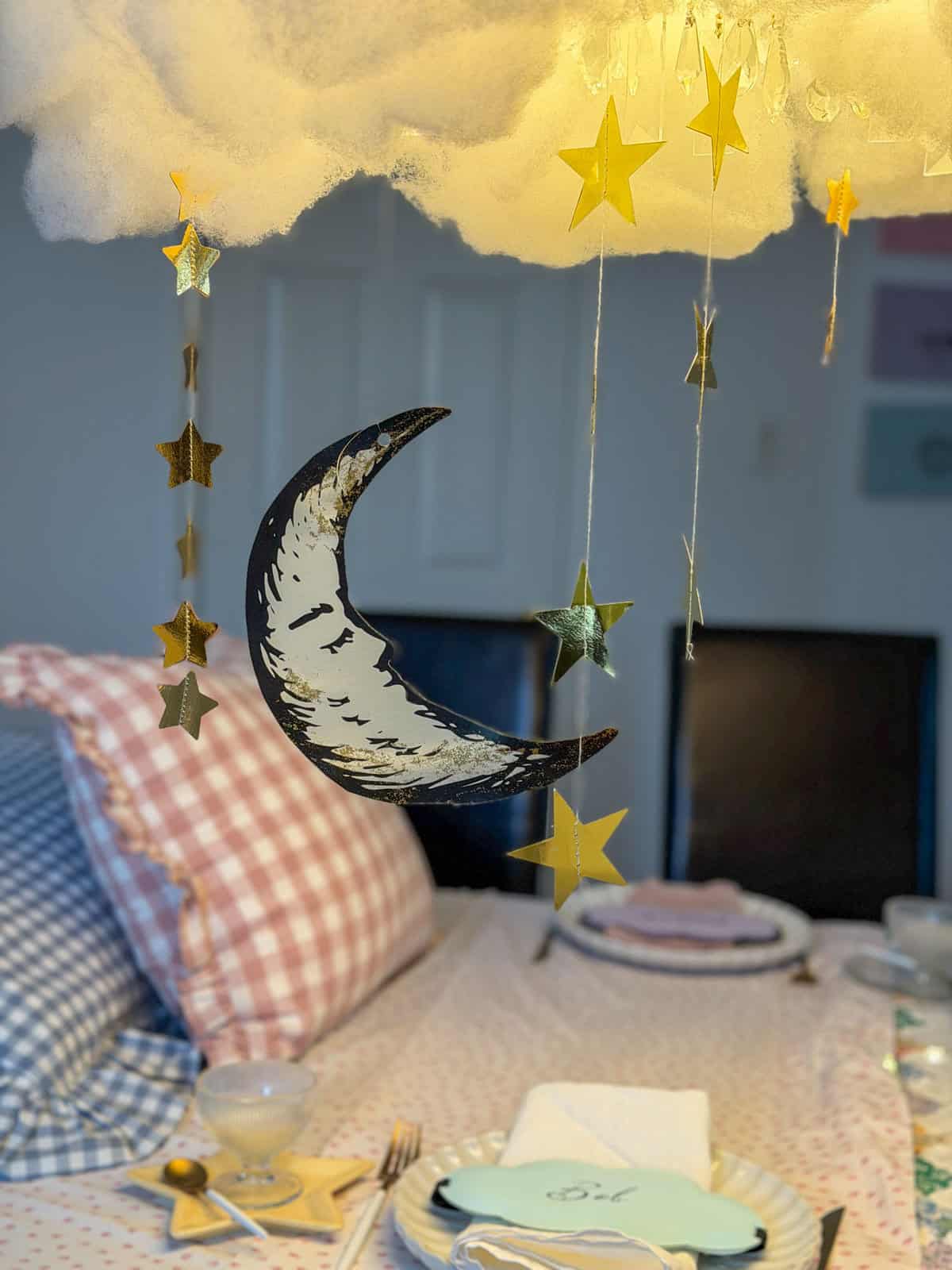 Crescent moon ornament hanging from a DIY cloud installation with gold star garlands and warm candlelight below.