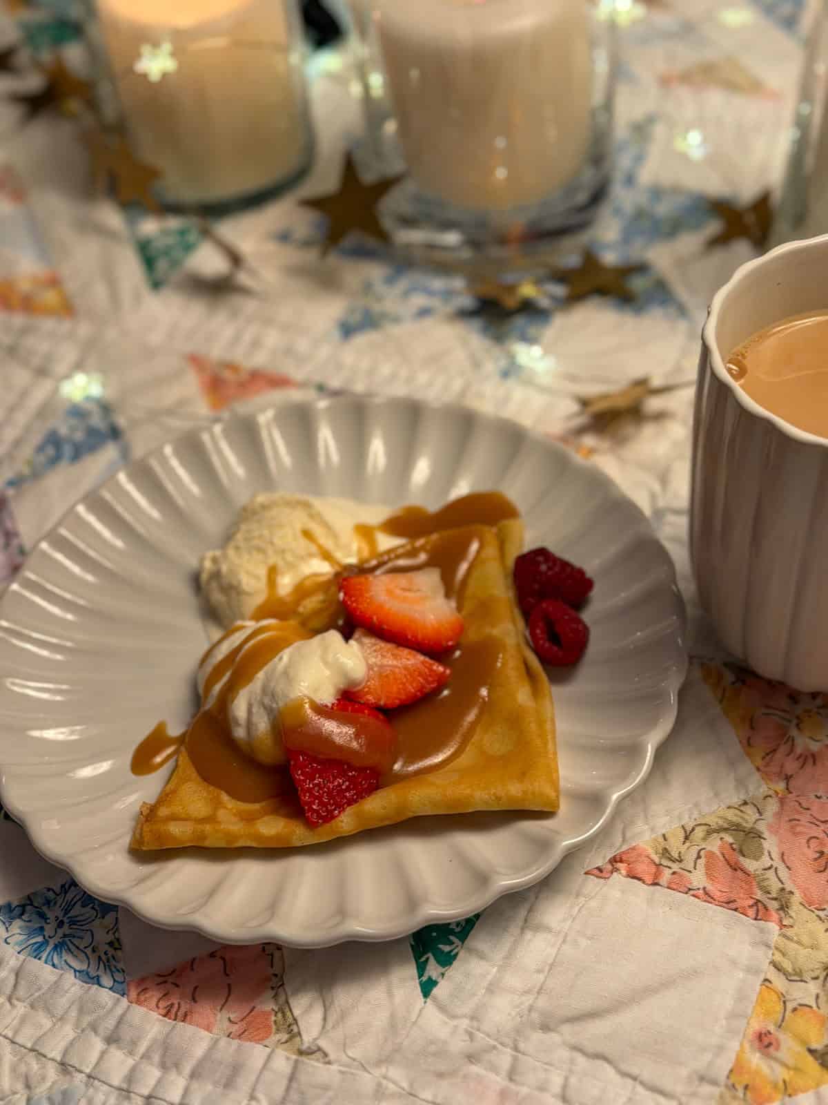 Vanilla crepe filled with whipped cream, fresh strawberries, raspberries, and caramel drizzle on a white scalloped plate.