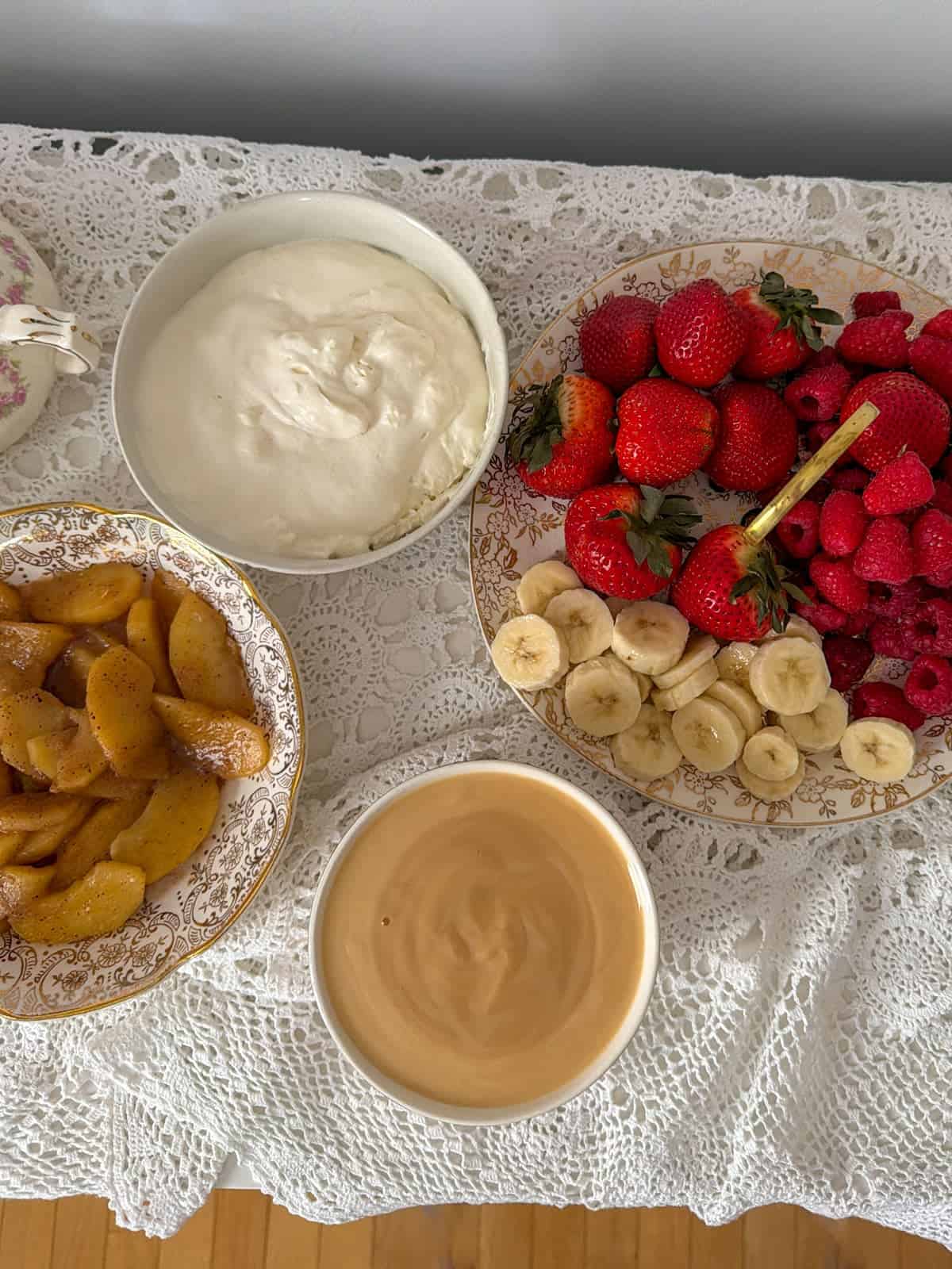 Crepe bar toppings including sauteed cinnamon apples, fresh strawberries, raspberries, banana slices, whipped cream, and caramel sauce on a white lace tablecloth.