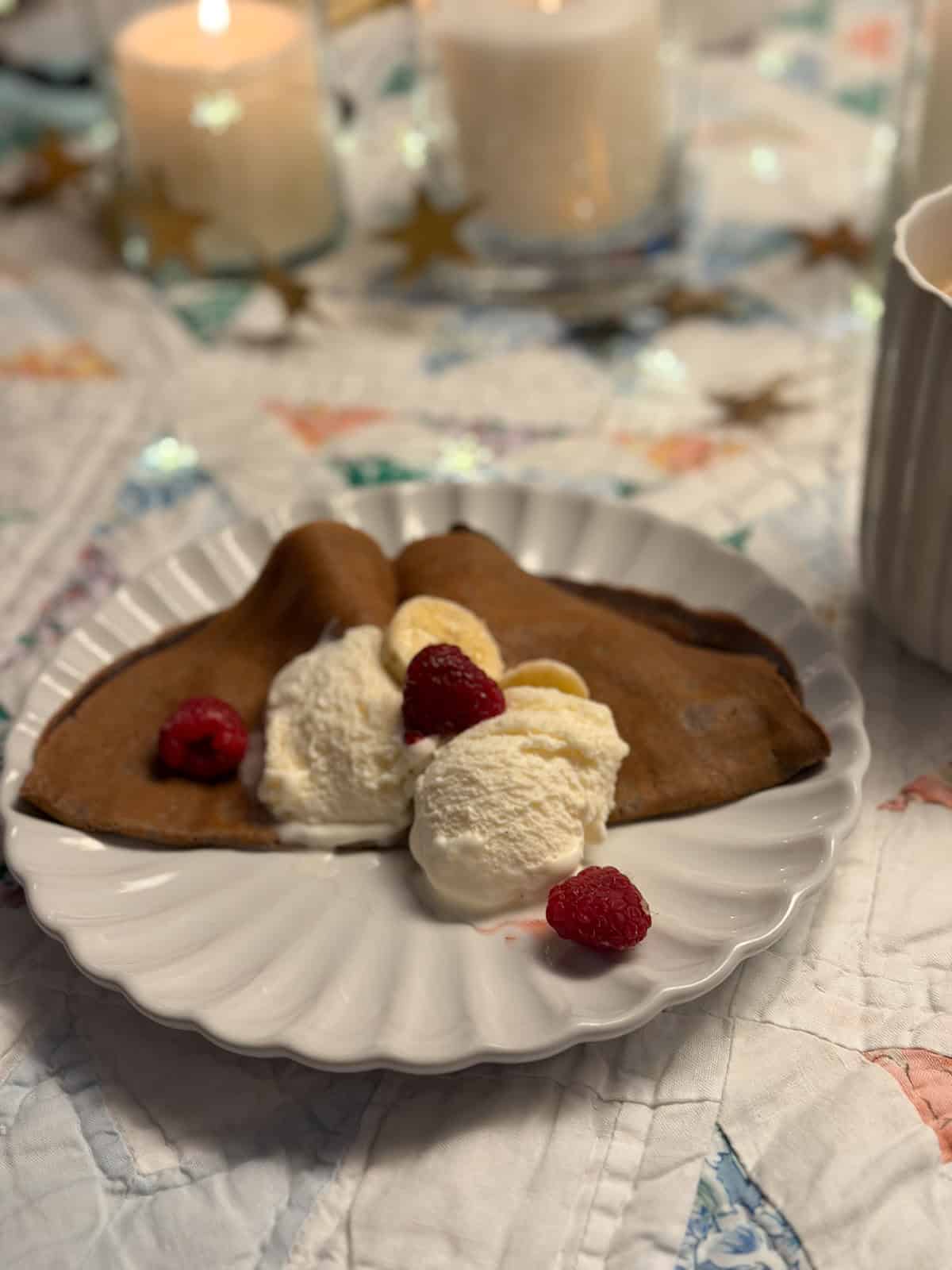 Chocolate crepe with whipped cream, fresh raspberries, and banana slices on a white scalloped dinner plate.