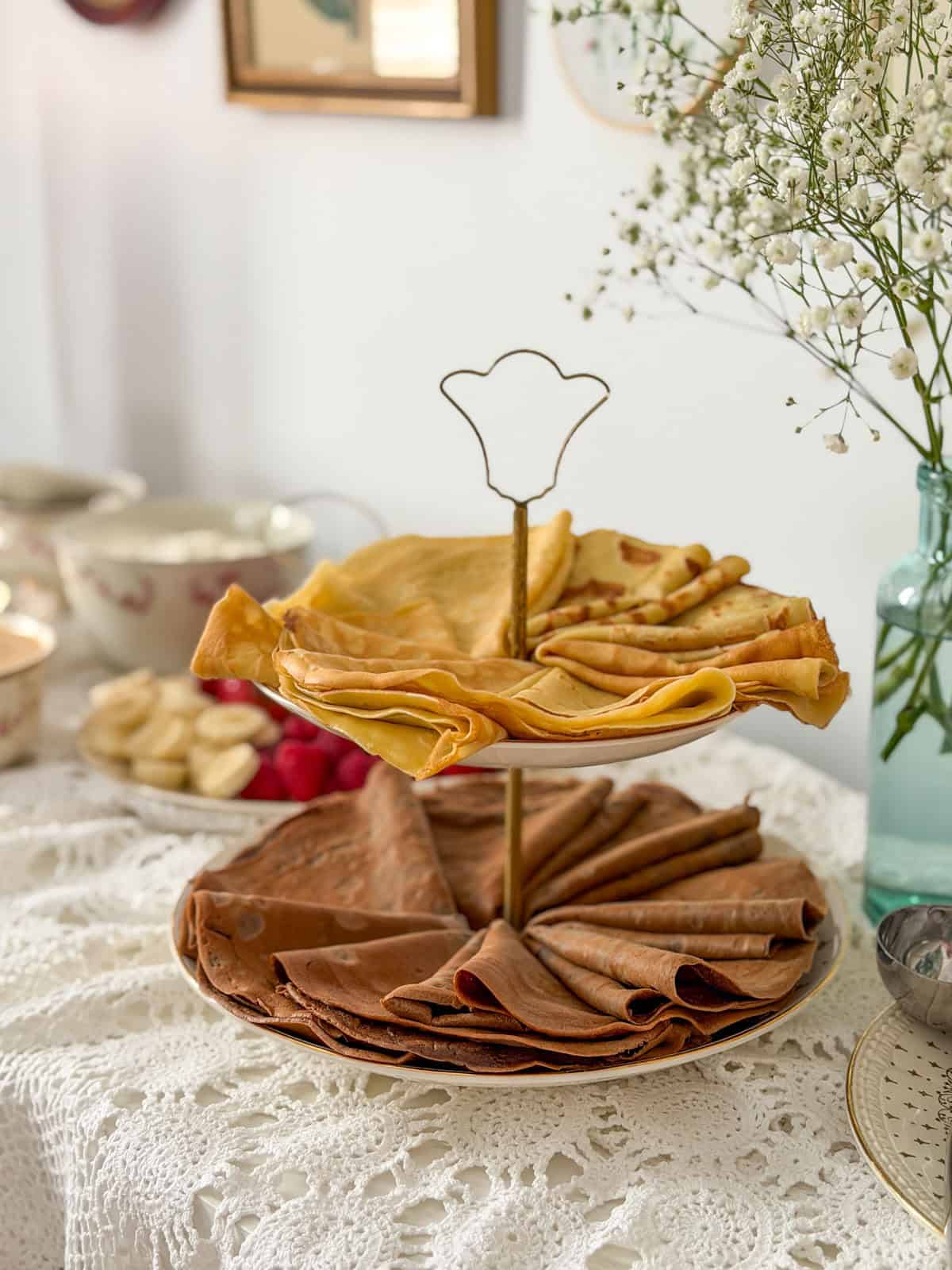 Two tier brass stand with vanilla crepes on the top tier and chocolate crepes on the bottom tier with baby's breath in a teal glass vase.