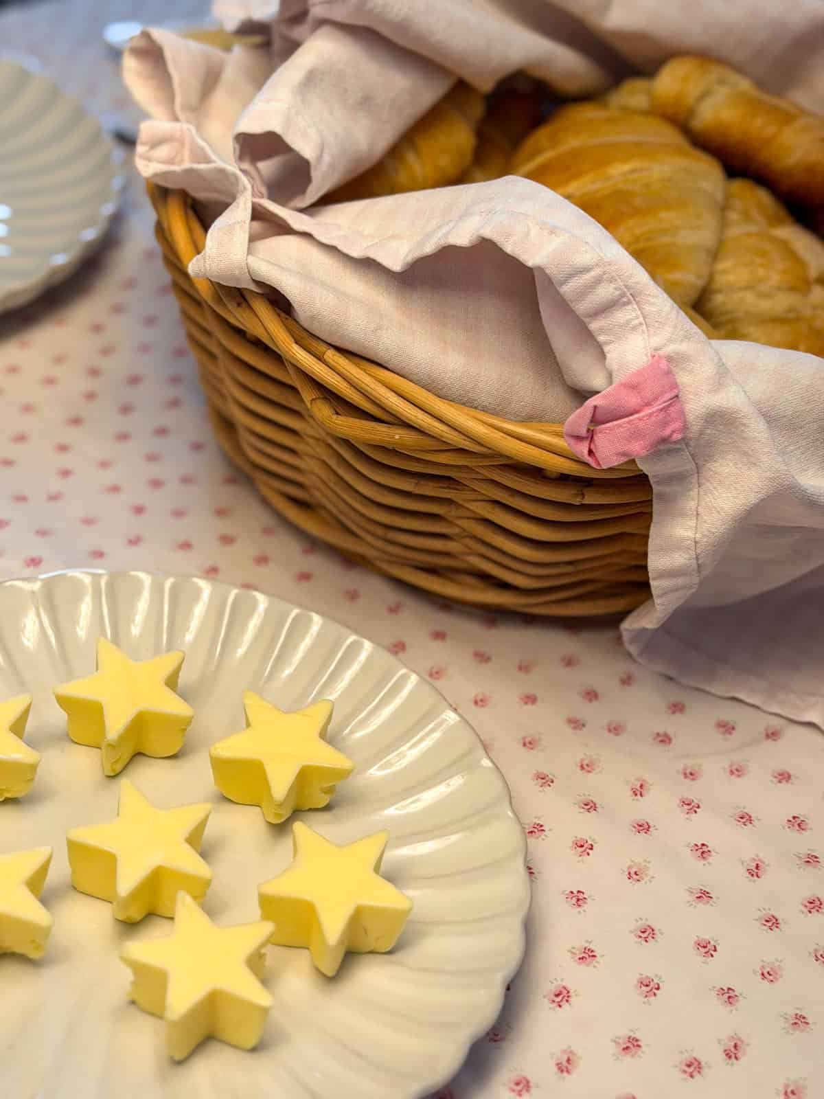 Star shaped butter pats on a white scalloped plate beside a wicker basket of fresh baked croissants.