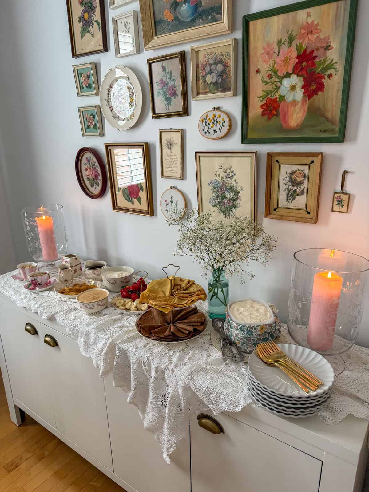 Breakfast for dinner party crepe bar on a buffet sideboard with vintage floral gallery wall, two tier crepe stand, fresh fruit, toppings, and lit pink pillar candles.