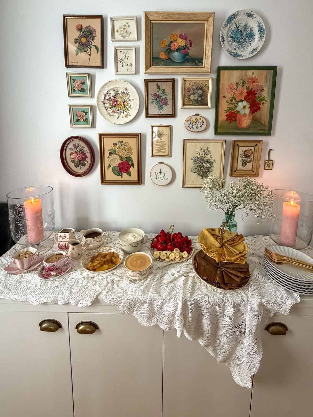 Crepe bar on a buffet sideboard with a two tier crepe stand, fresh fruit, caramel sauce, whipped cream, and lit pink pillar candles against a floral gallery wall.