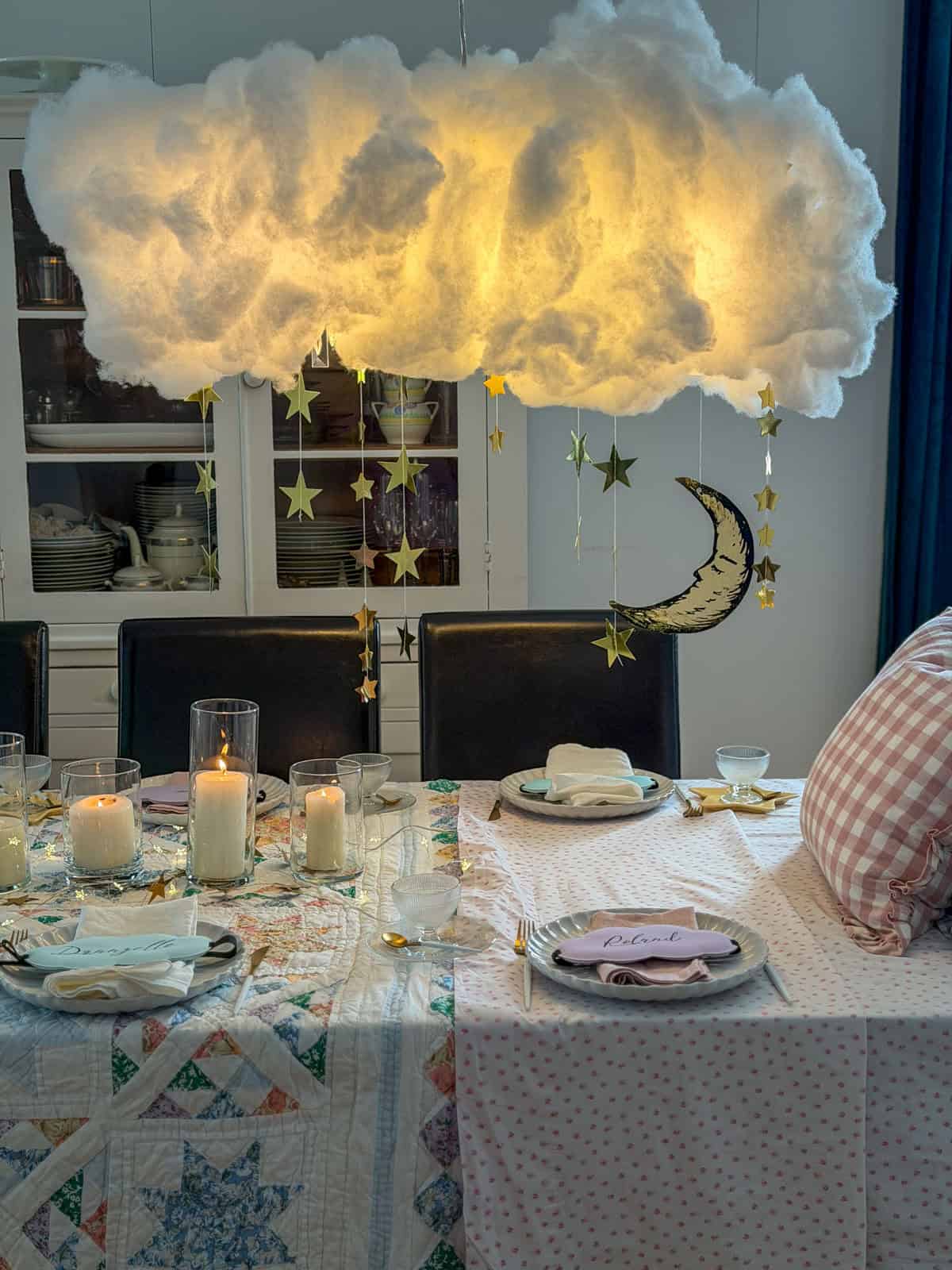 DIY polyfill cloud light installation with gold star garlands and crescent moon hanging over a candlelit breakfast for dinner party table with gingham pillow headboard.
