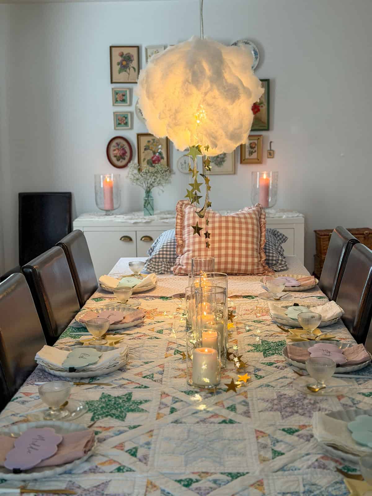 Breakfast for dinner party dining table viewed from the pillow end with glowing cloud installation overhead, gold star garlands, lit pillar candles, and personalized sleep mask place cards.