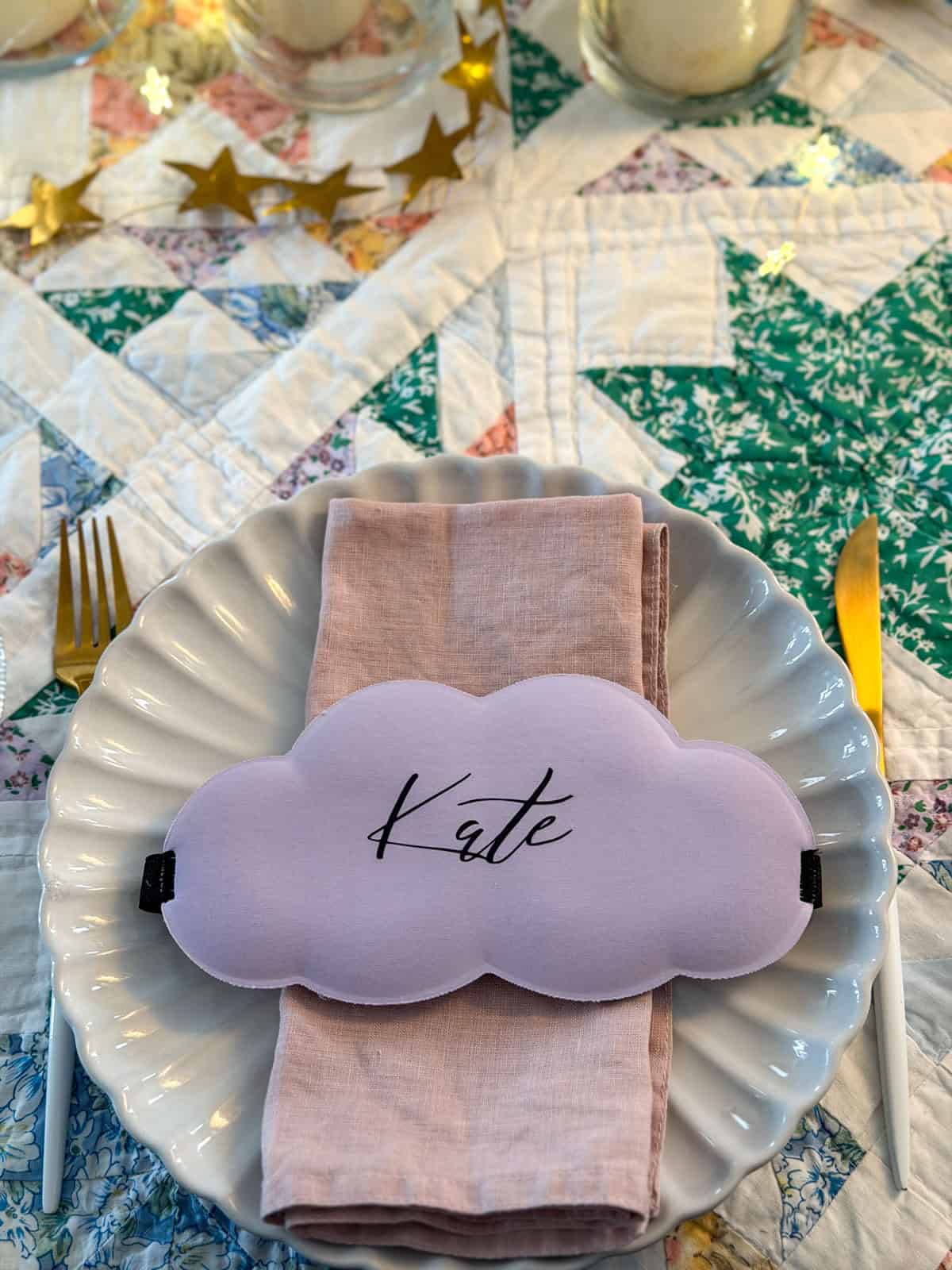 Lavender cloud shaped sleep mask with the name Kate personalized in iron-on vinyl on a white scalloped dinner plate with a pink linen napkin.
