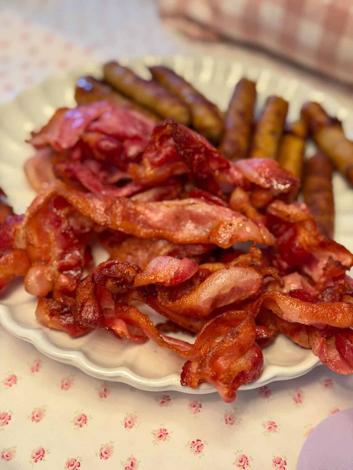 Crispy bacon strips and breakfast sausages piled on a white scalloped plate with a pink floral tablecloth.