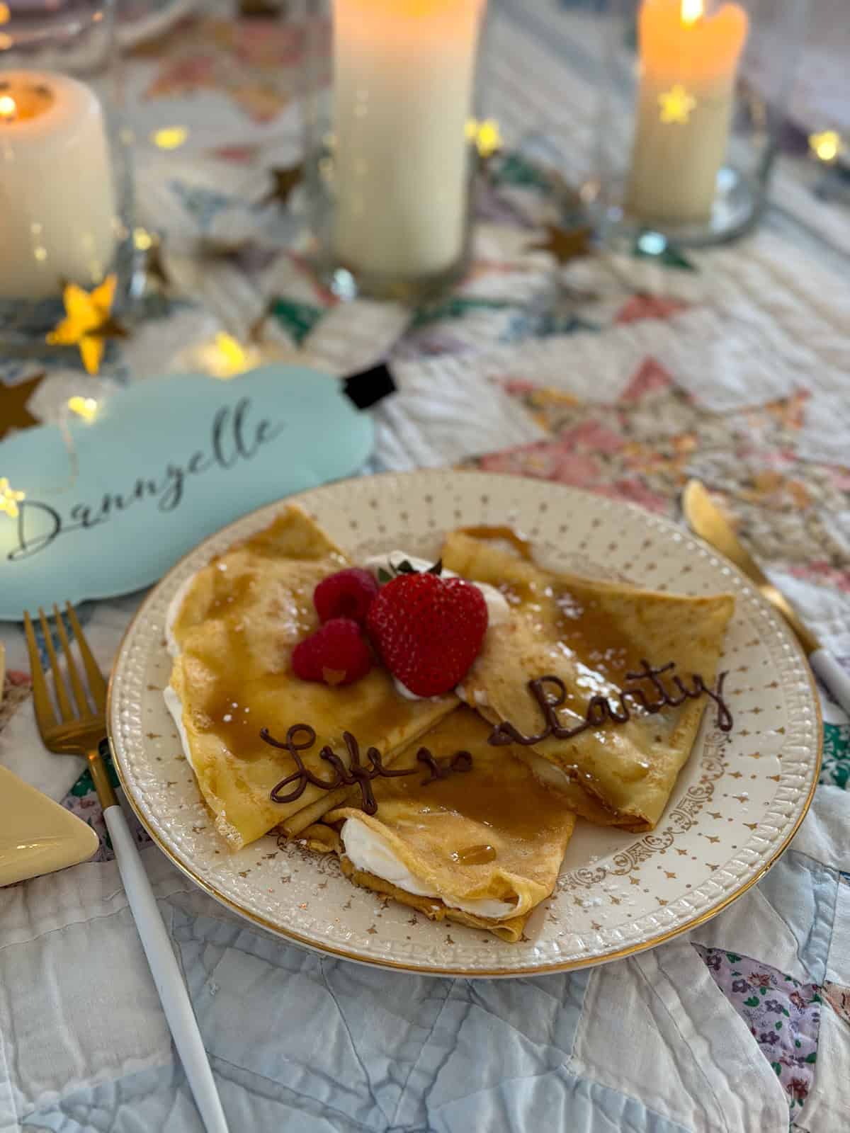 Vanilla crepe with fresh strawberries, raspberries, whipped cream, and caramel drizzle with Life is a Party written in chocolate sauce on a gold rimmed plate.