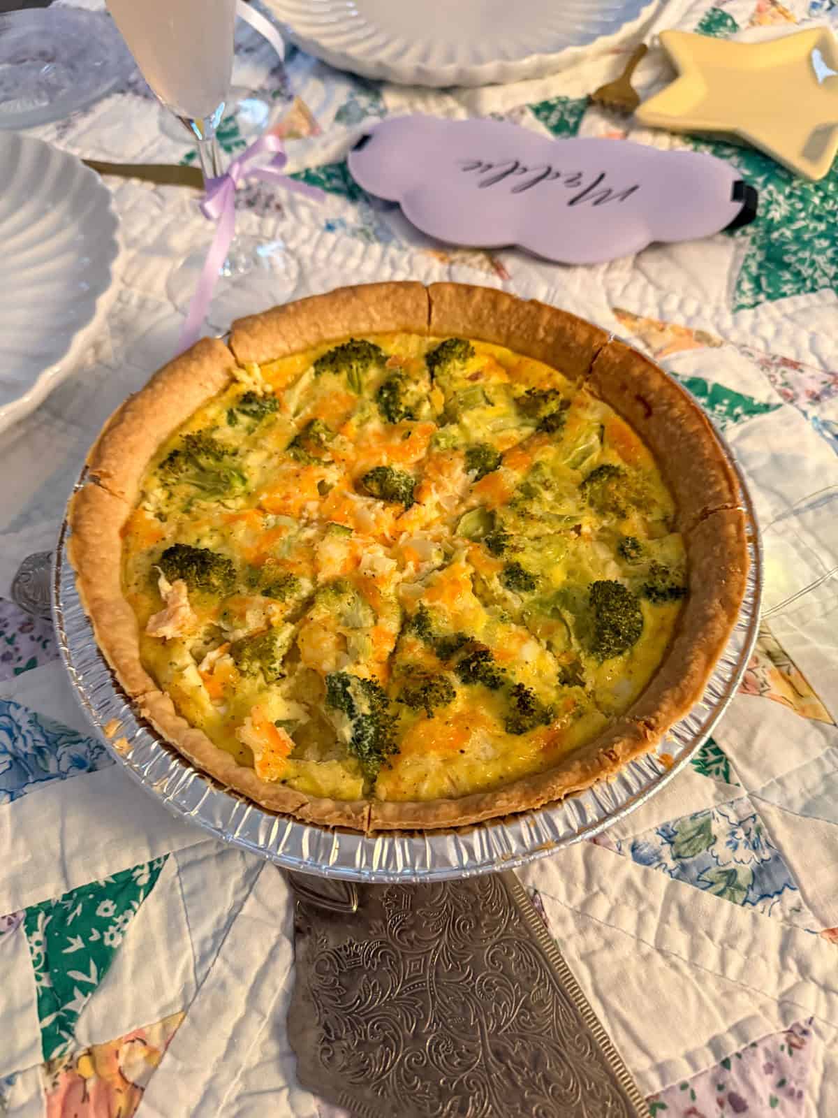 Broccoli cheddar quiche in a pie dish on a glass cake stand at a breakfast for dinner party table with sleep mask place card in the background.