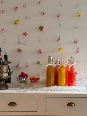 Complete mimosa bar setup on white console table with vintage silver ice bucket, labeled colorful juice bottles, small glass garnish bowls, and ribbon-tied champagne flutes on white lace tablecloth with flower wall backdrop.