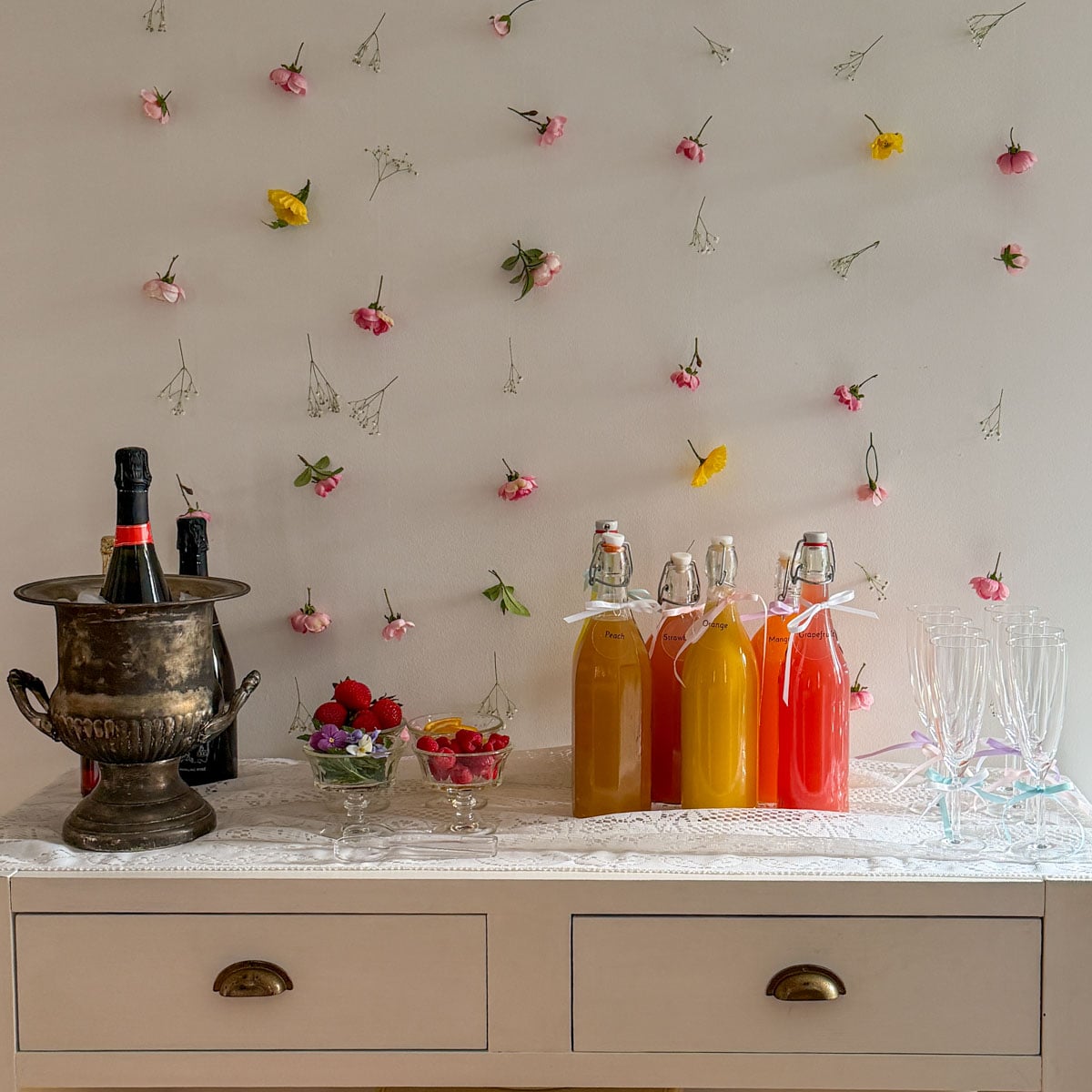 Complete mimosa bar setup on white console table with vintage silver ice bucket, labeled colorful juice bottles, small glass garnish bowls, and ribbon-tied champagne flutes on white lace tablecloth with flower wall backdrop.
