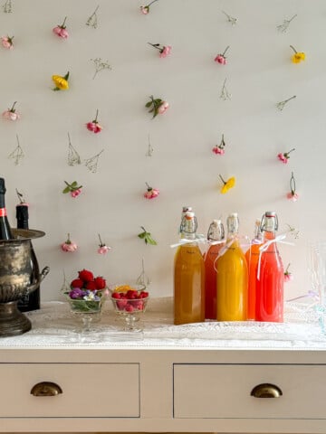 Complete mimosa bar setup on white console table with vintage silver ice bucket, labeled colorful juice bottles, small glass garnish bowls, and ribbon-tied champagne flutes on white lace tablecloth with flower wall backdrop.