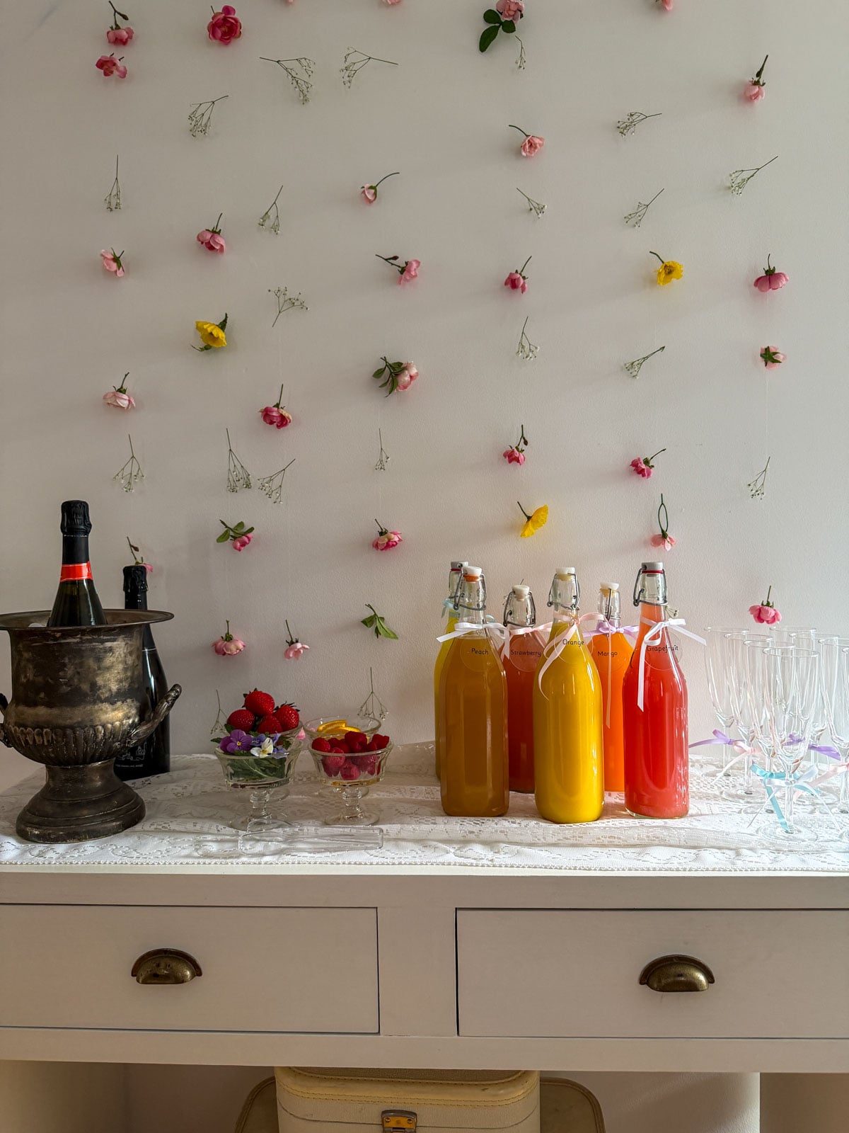 Mimosa bar on white console table with silver ice bucket, colorful juice bottles, garnish bowls, and champagne flutes with pastel ribbon bows against a floating flower wall backdrop.
