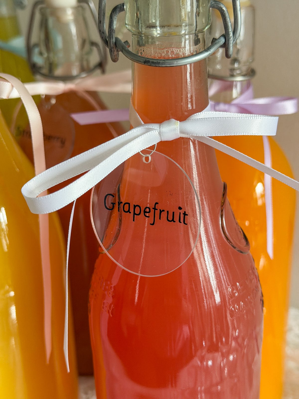 Swing-top glass bottle filled with bright pink grapefruit juice, clear acrylic Grapefruit label and white ribbon bow tied at the neck, orange and mango juice bottles visible in background.