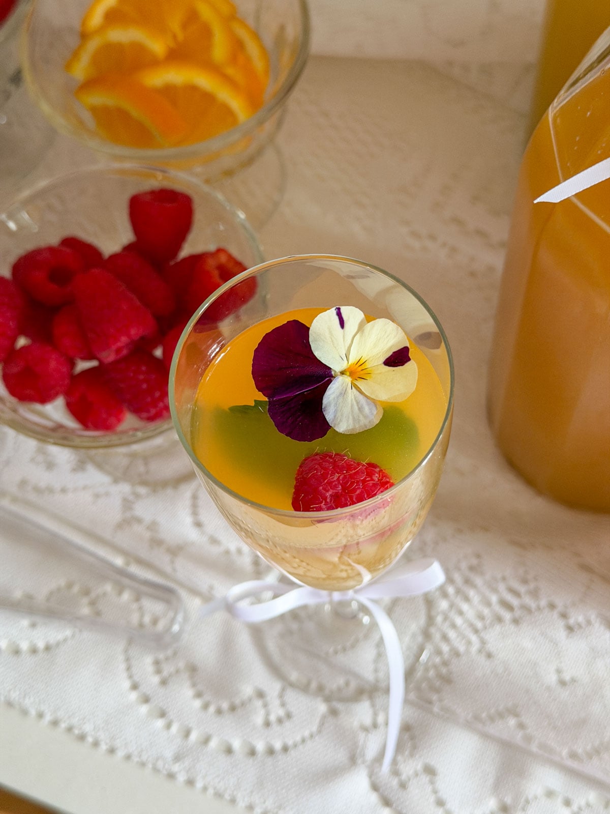Champagne flute with mimosa viewed from above showing a floating bicolor pansy and fresh raspberry in golden sparkling juice, bowl of raspberries and juice bottle on white lace tablecloth.
