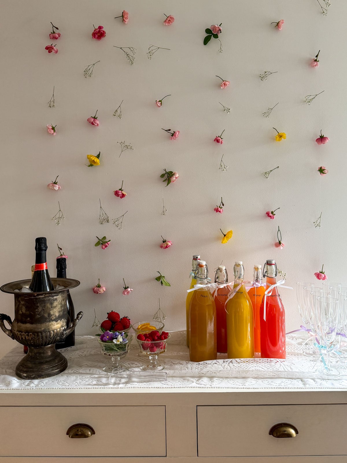 Complete mimosa bar setup on white console table with vintage silver ice bucket, labeled colorful juice bottles, small glass garnish bowls, and ribbon-tied champagne flutes on white lace tablecloth with flower wall backdrop.