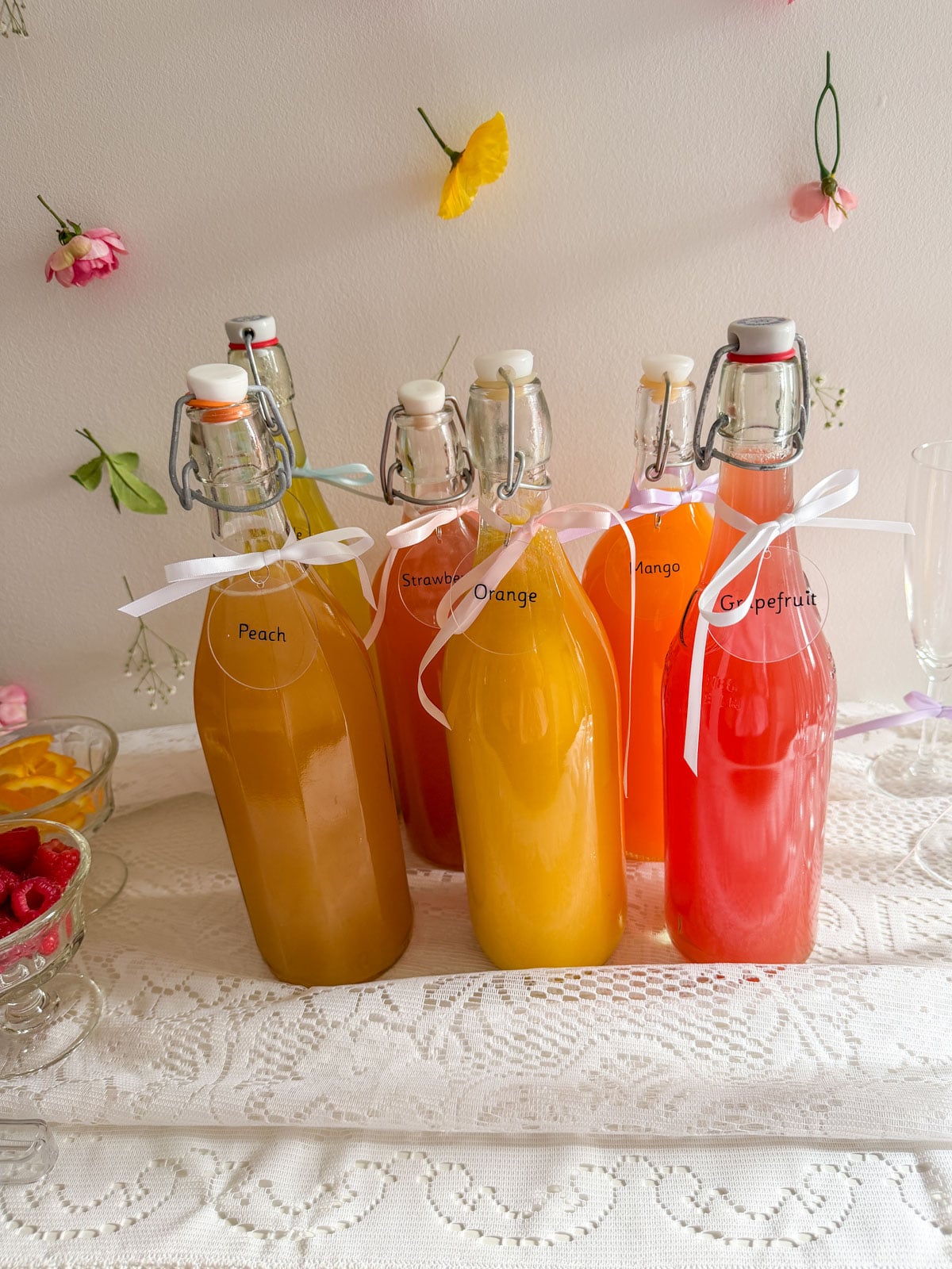 Six labeled swing-top glass juice bottles in peach, strawberry, orange, mango, and grapefruit arranged on a lace tablecloth with pink and yellow flowers floating on the wall behind them.