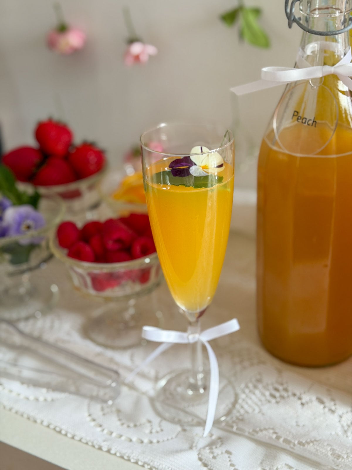 Champagne flute filled with orange mimosa topped with a floating edible pansy flower, white ribbon bow on stem, peach juice bottle on lace tablecloth in background.