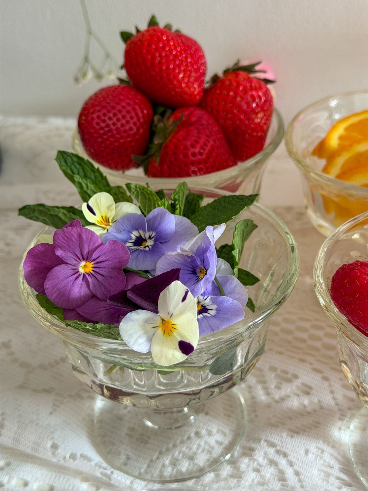 Glass pedestal bowl filled with fresh edible pansy flowers in purple, blue, and cream nestled in bright green mint leaves, bowl of red strawberries visible in the background.