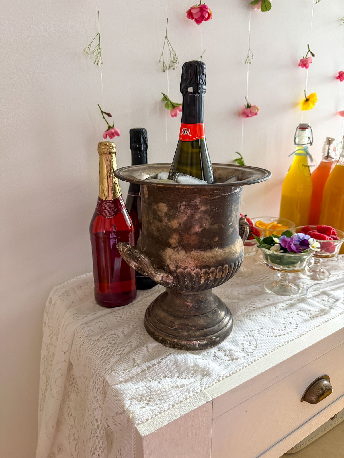 Tarnished silver ice bucket with chilled prosecco bottle surrounded by ice, non-alcoholic sparkling bottles and colorful juice lineup on lace tablecloth with flower wall backdrop.