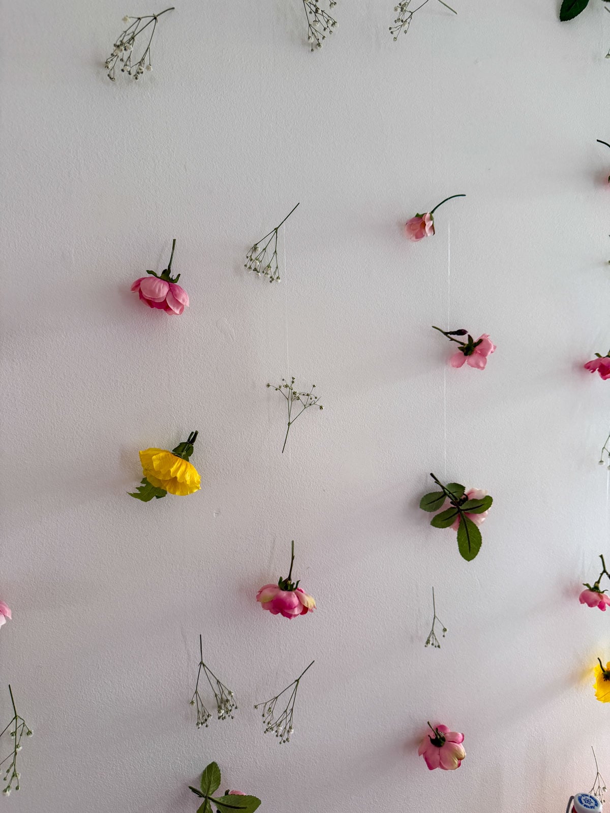 DIY floating flower curtain backdrop with pink and cream faux roses, yellow blooms, and dried baby's breath sprigs on clear fishing line against a white wall.