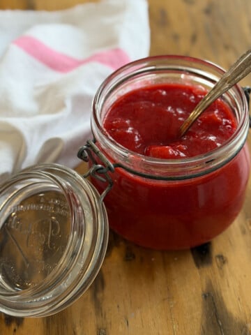 Open glass Kilner jar filled with bright red strawberry rhubarb compote with a gold vintage spoon resting inside, on a wooden surface with a pink and white striped towel alongside.