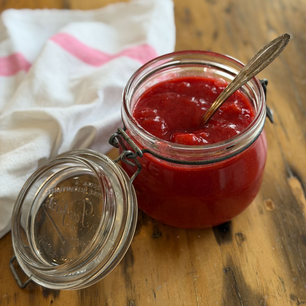 Open glass Kilner jar filled with bright red strawberry rhubarb compote with a gold vintage spoon resting inside, on a wooden surface with a pink and white striped towel alongside.
