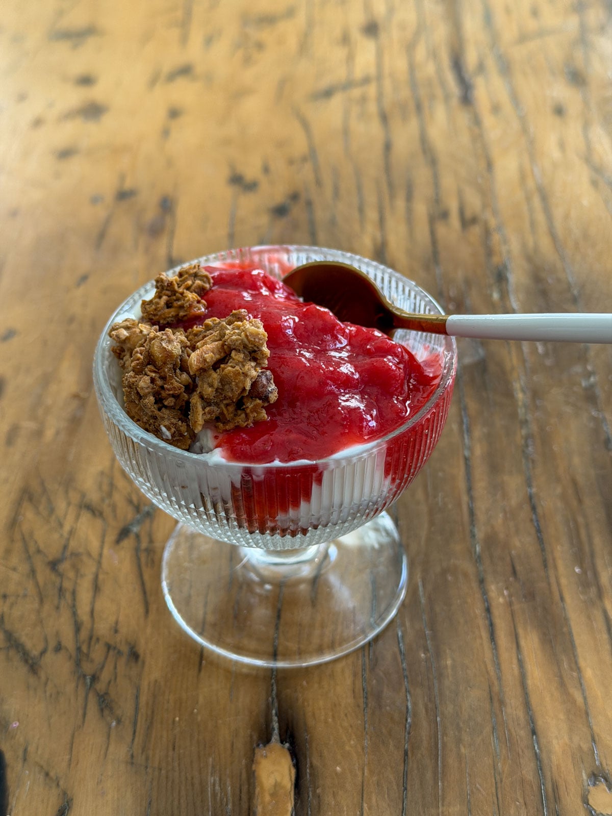 Strawberry rhubarb compote spooned over vanilla Greek yogurt and granola in a ribbed footed glass dessert dish with a gold spoon.