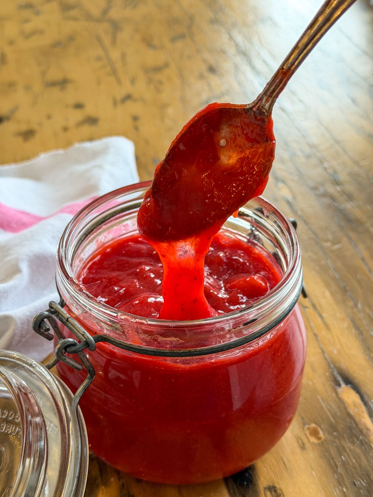 A vintage spoon drizzling a long stream of glossy strawberry rhubarb compote back into an open glass Kilner jar on a wooden surface.