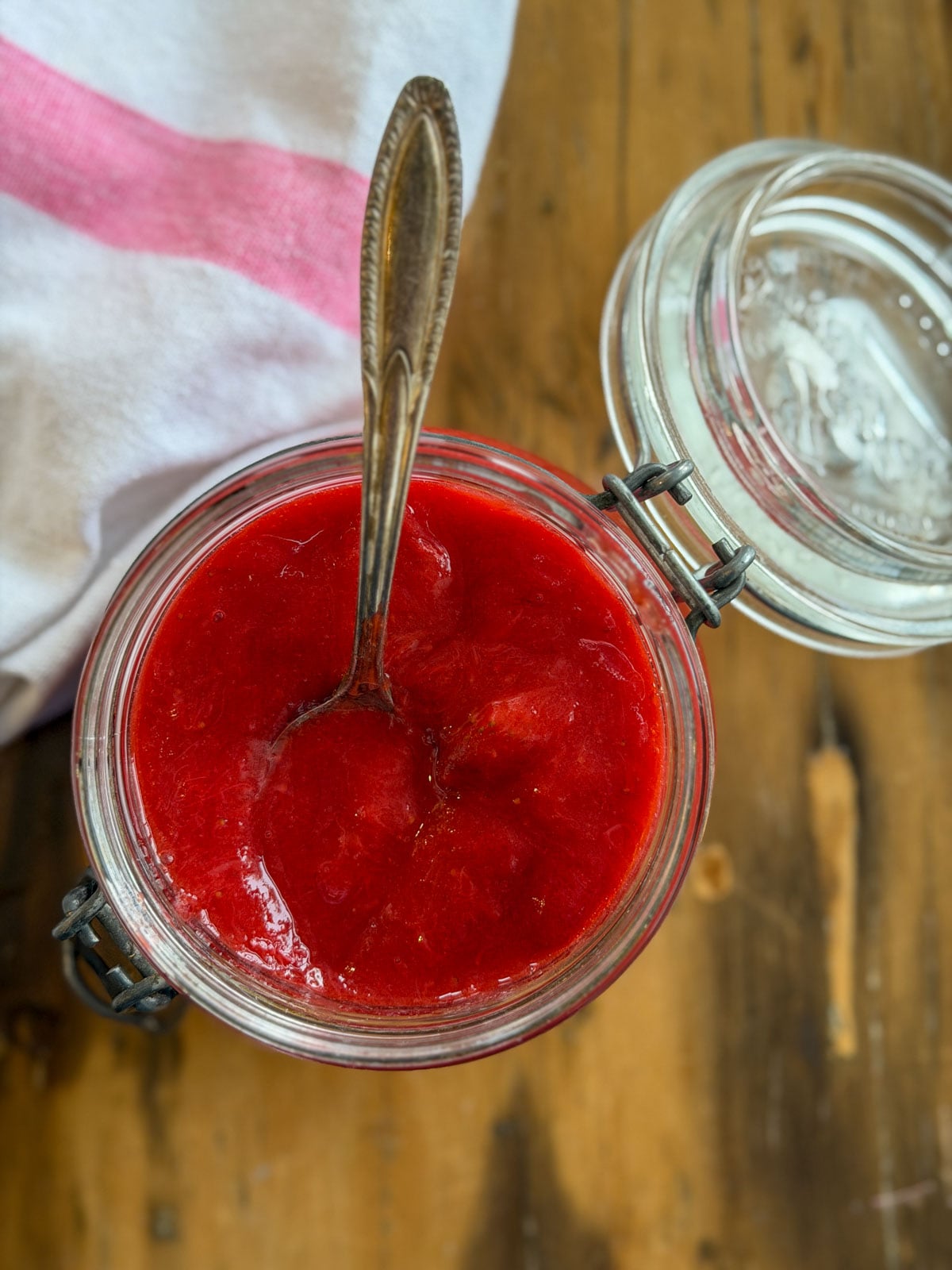 Strawberry rhubarb compote filling an open glass Kilner jar with a vintage spoon resting inside, on a wooden surface with a pink and white striped towel and jar lid alongside.