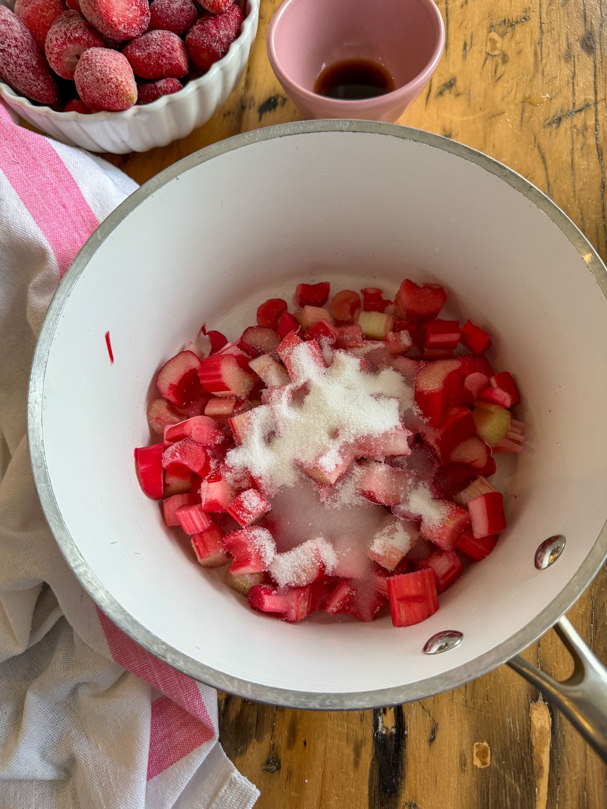 Chopped rhubarb and granulated sugar combined in a white saucepan before cooking, with a bowl of frozen strawberries and a small pink cup of vanilla extract nearby.