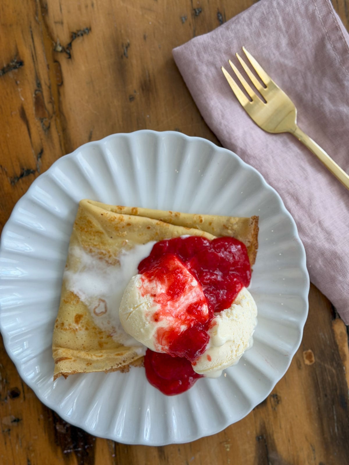 Sweet crepe folded on a white scalloped plate topped with a scoop of vanilla ice cream and bright red strawberry rhubarb compote, with a pink linen napkin and gold fork alongside.