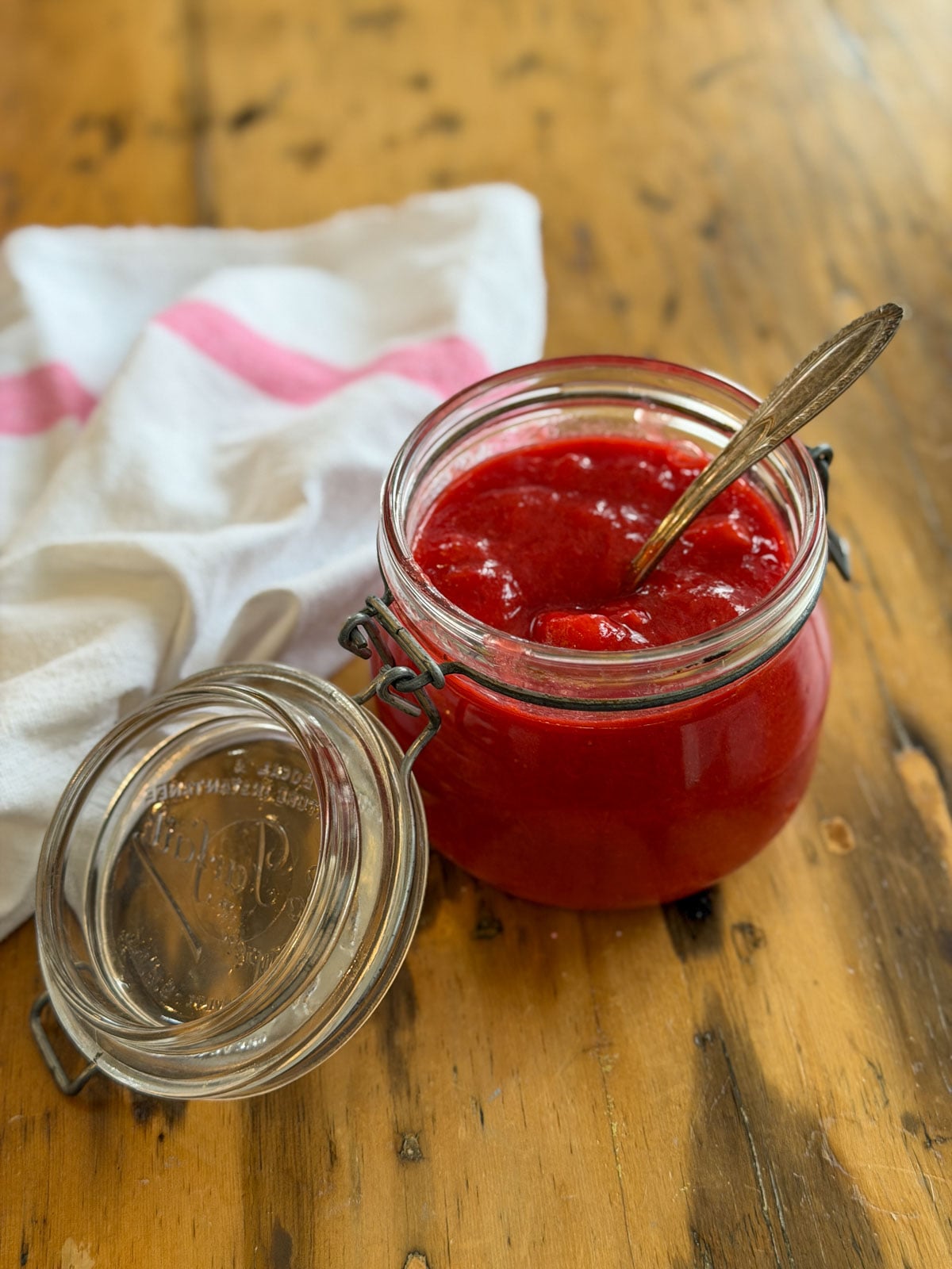 Open glass Kilner jar filled with bright red strawberry rhubarb compote with a gold vintage spoon resting inside, on a wooden surface with a pink and white striped towel alongside.