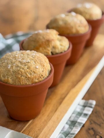 Four individual herb bread loaves baked in terracotta flower pots on a wooden serving board with flaky sea salt on top.