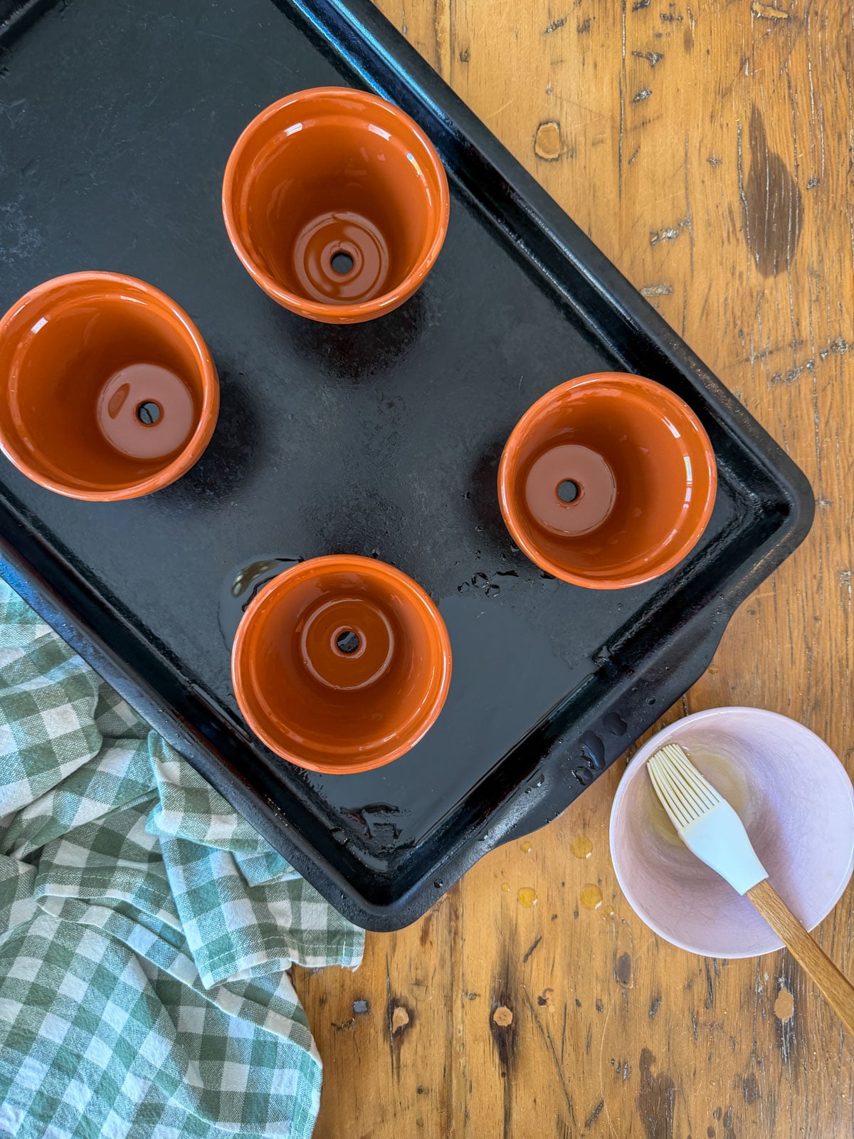 Four seasoned terracotta pots on a baking sheet with oil and a pastry brush.