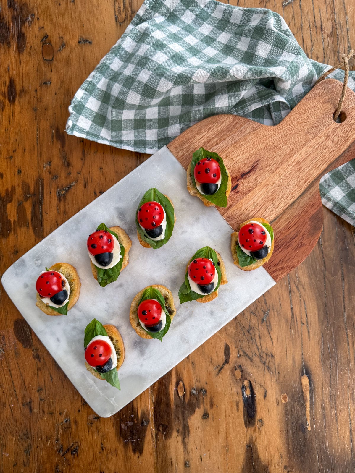 Ladybug caprese bruschetta arranged on a marble board beside a green checked napkin.