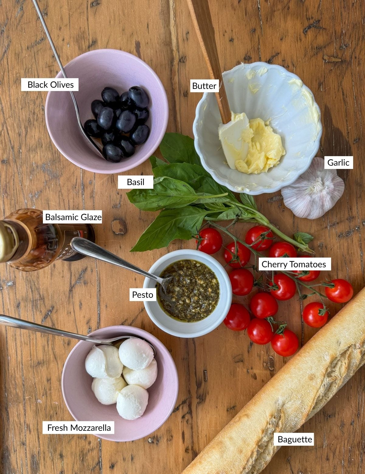 Ingredients for ladybug caprese bruschetta including cherry tomatoes, mozzarella, olives, basil, pesto, butter, and a baguette.