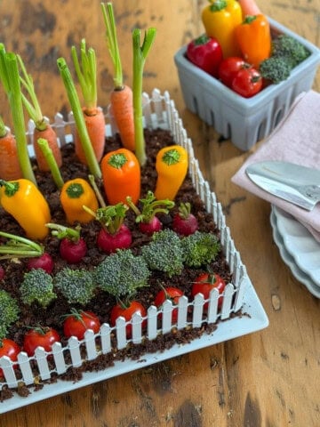 Vegetable garden hummus dip with white picket fence on a wood table styled with a grey berry basket of extra vegetables, pink linen napkins, and a small garden trowel.