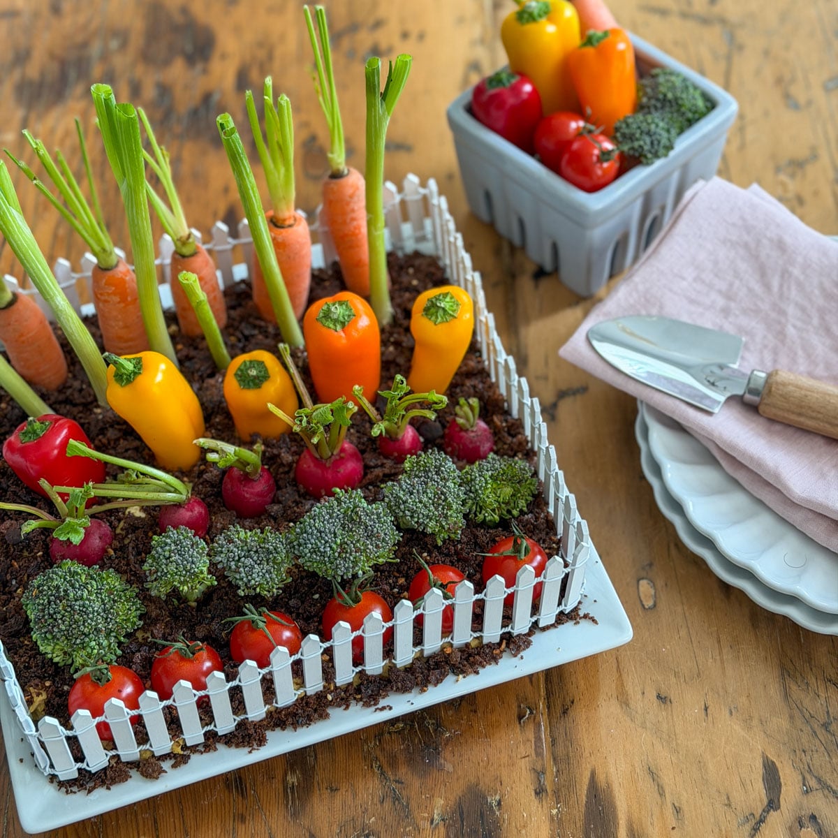 Vegetable garden hummus dip with white picket fence on a wood table styled with a grey berry basket of extra vegetables, pink linen napkins, and a small garden trowel.