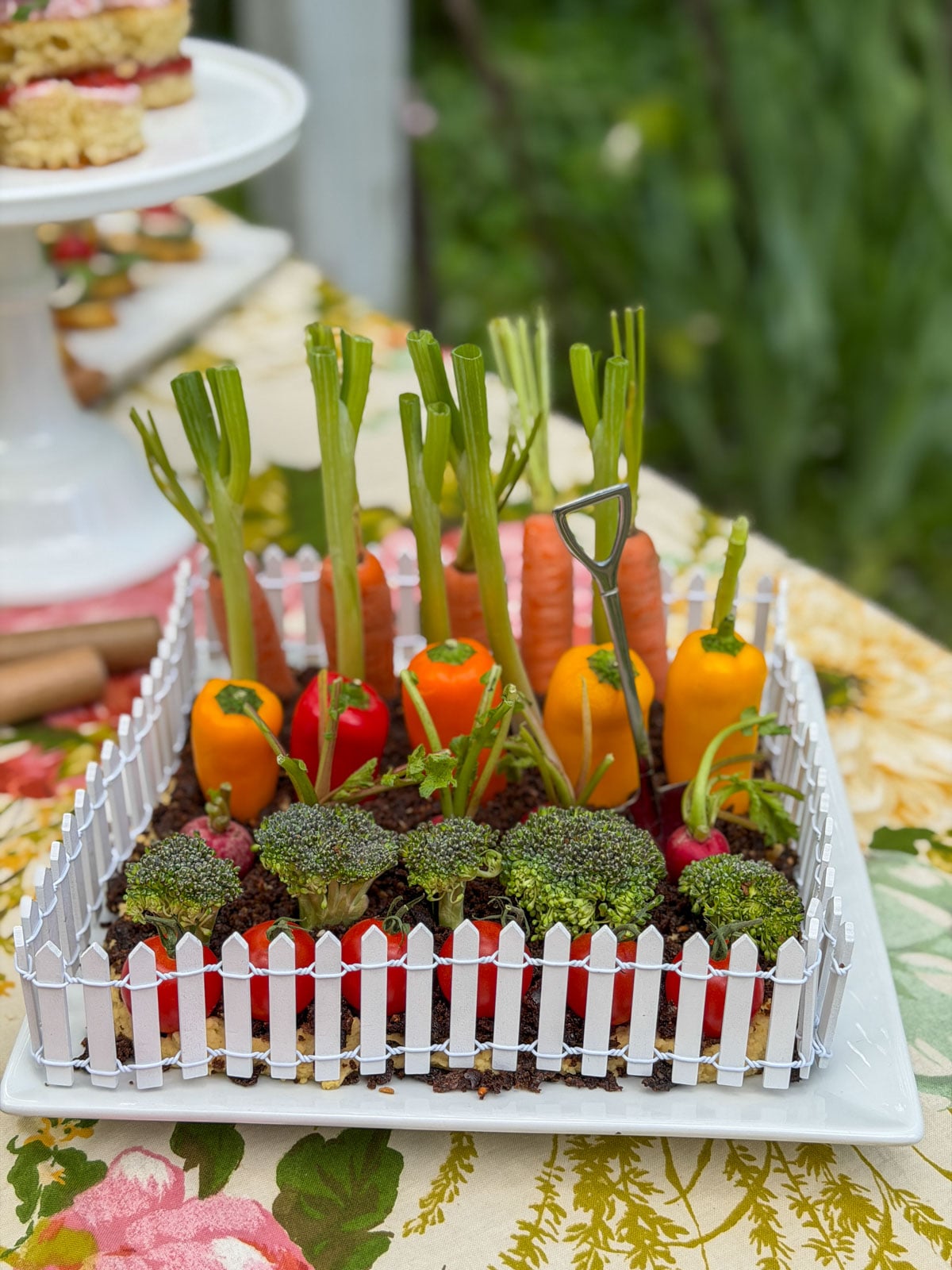 Vegetable garden hummus dip on a white platter surrounded by a miniature picket fence with fresh vegetables standing upright like a growing garden.
