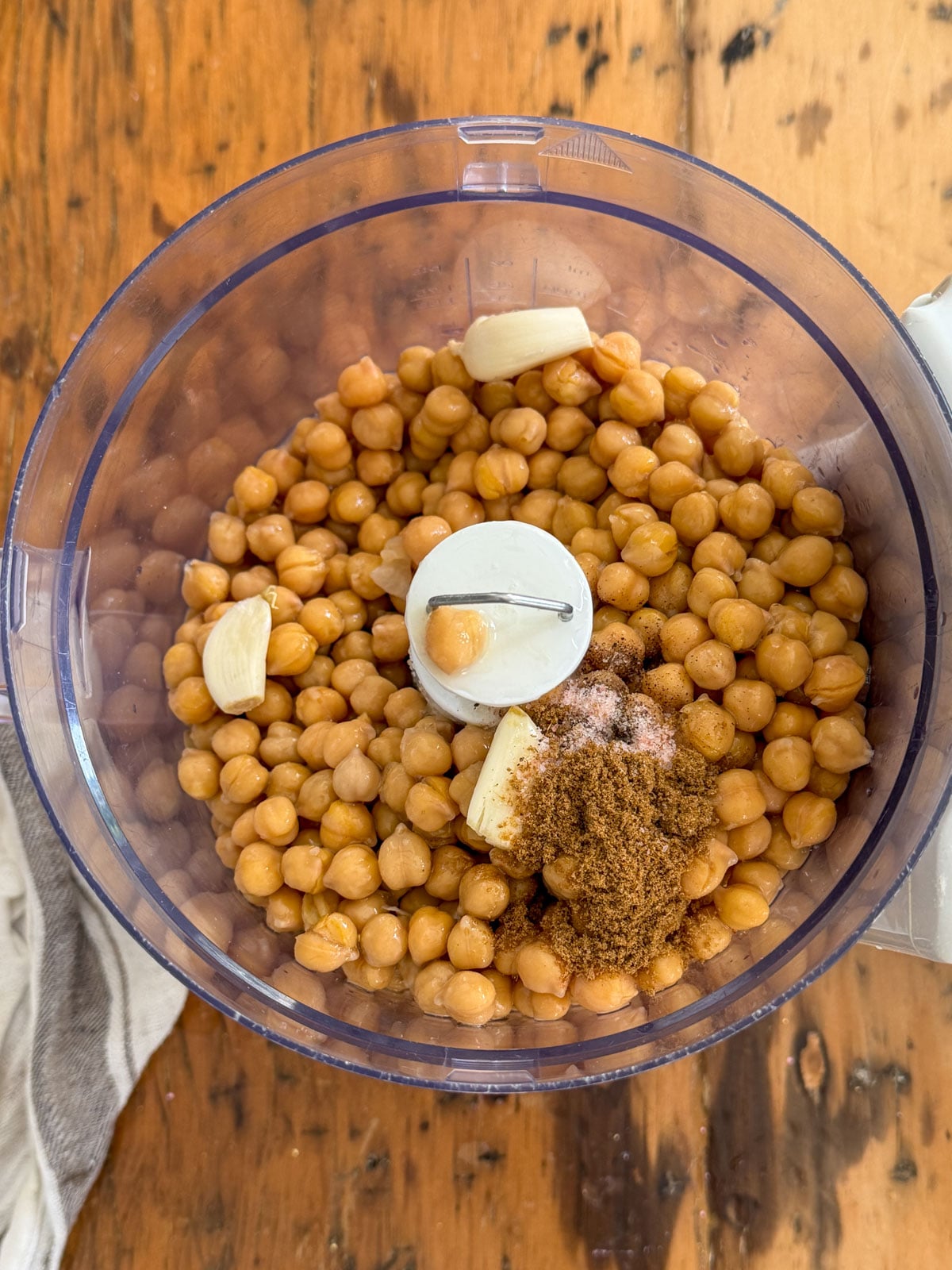 Chickpeas, garlic cloves, cumin, salt, and pepper added to a food processor bowl ready to blend into homemade hummus.