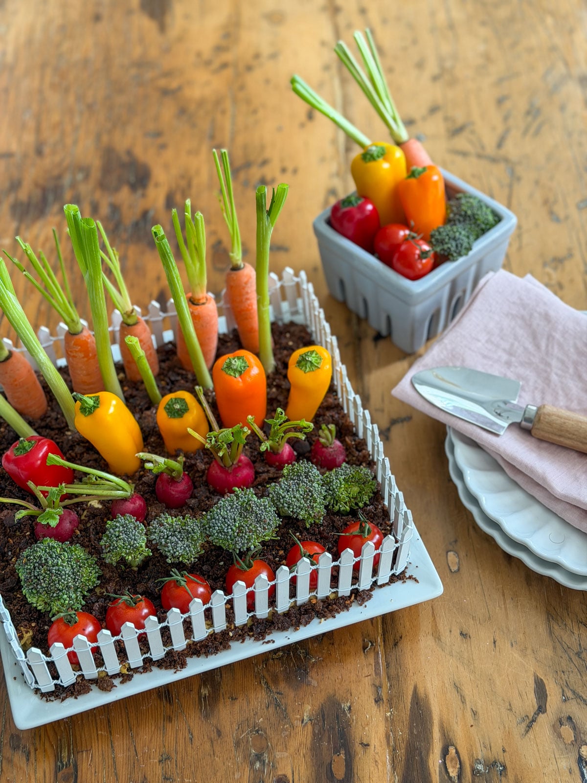 Vegetable garden hummus dip on a wood table styled with a grey berry basket of extra vegetables and a small garden trowel alongside pink linen napkins.