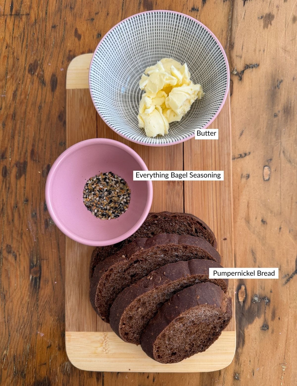 Sliced pumpernickel bread on a cutting board with a bowl of butter and a bowl of everything bagel seasoning for making hummus soil topping.