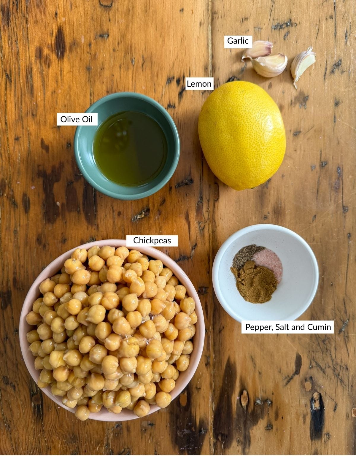 Hummus ingredients flat lay on a wood table including a bowl of chickpeas, olive oil, a lemon, garlic cloves, and a bowl of cumin salt and pepper.