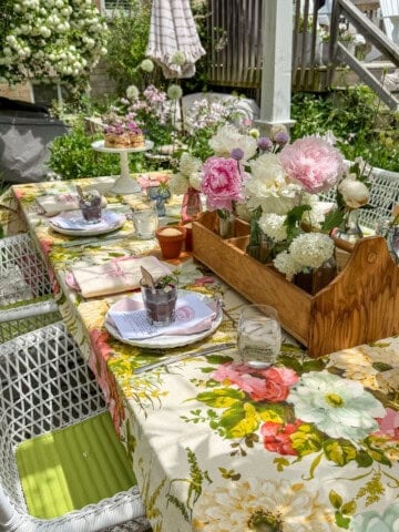 Garden party table set under white pergola with vintage floral tablecloth, pink peony centerpiece in wooden tool caddy, white wicker chairs, and climbing white roses in background.